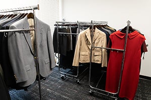 Clothing racks inside the University of Maryland’s Smith School Career Boutique display professional attire, including gray and beige blazers, black dress pants, and a red dress, neatly organized in a bright, carpeted room.