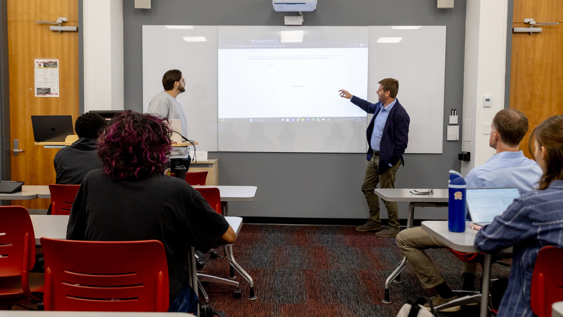 A University of Maryland classroom scene shows a professor pointing to a projected screen while another instructor stands nearby. Several students sit at desks with laptops and notebooks, attentively watching the presentation. The room features red chairs, gray walls, and wood-paneled doors.