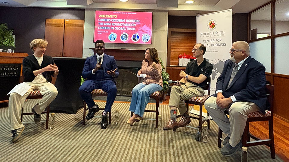 A student moderator and four panelists sit on chairs in front of an audience during the “Careers Crossing Borders: Mini Roundtable on Careers in Global Trade” event at the University of Maryland’s Robert H. Smith School of Business. The group is engaged in discussion, with microphones in hand, against a backdrop displaying event details and the Center for Global Business banner.