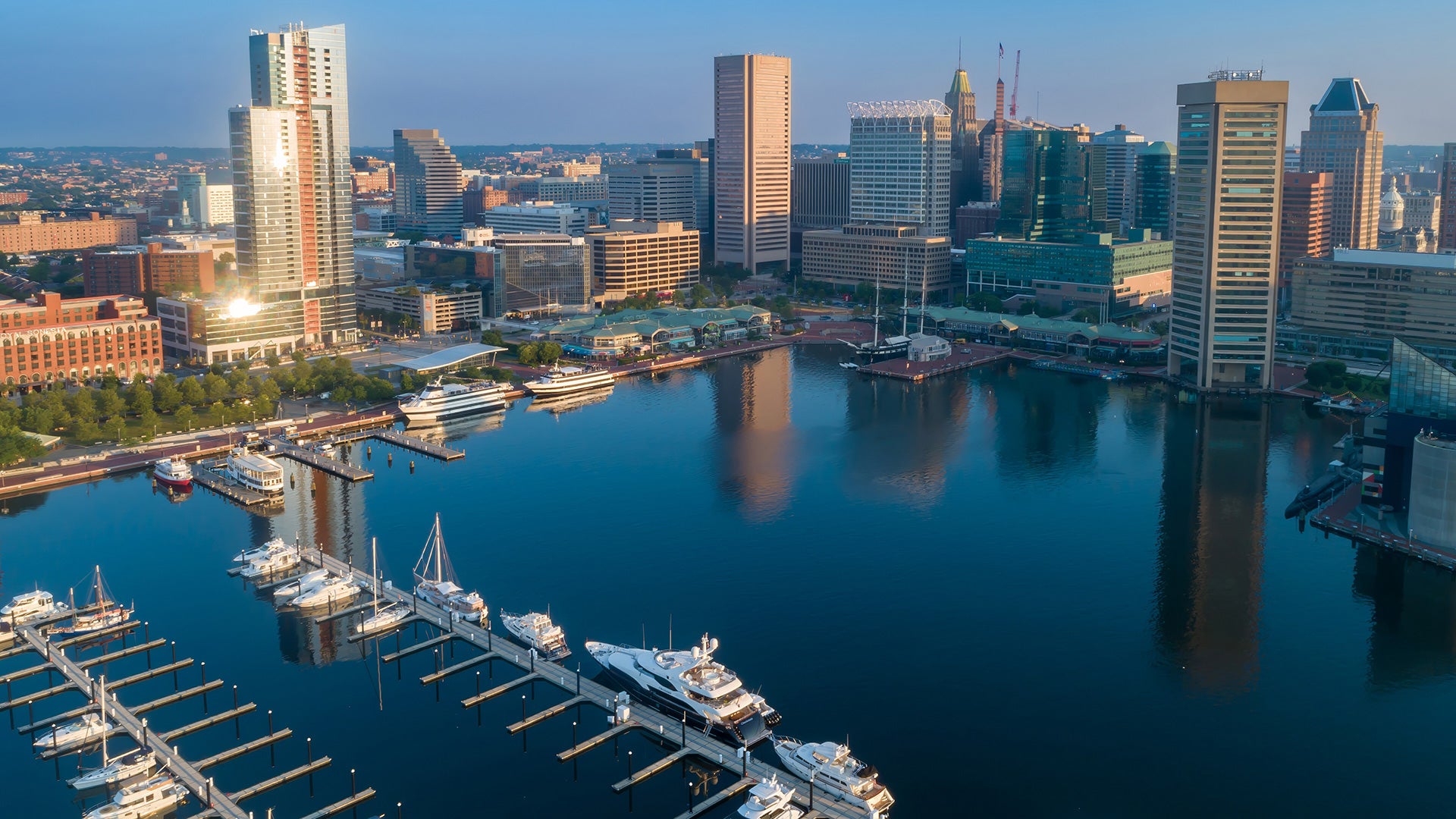 Aerial view of Baltimore’s Inner Harbor at sunrise, showing calm water reflecting downtown skyscrapers, moored boats and yachts, and waterfront buildings.