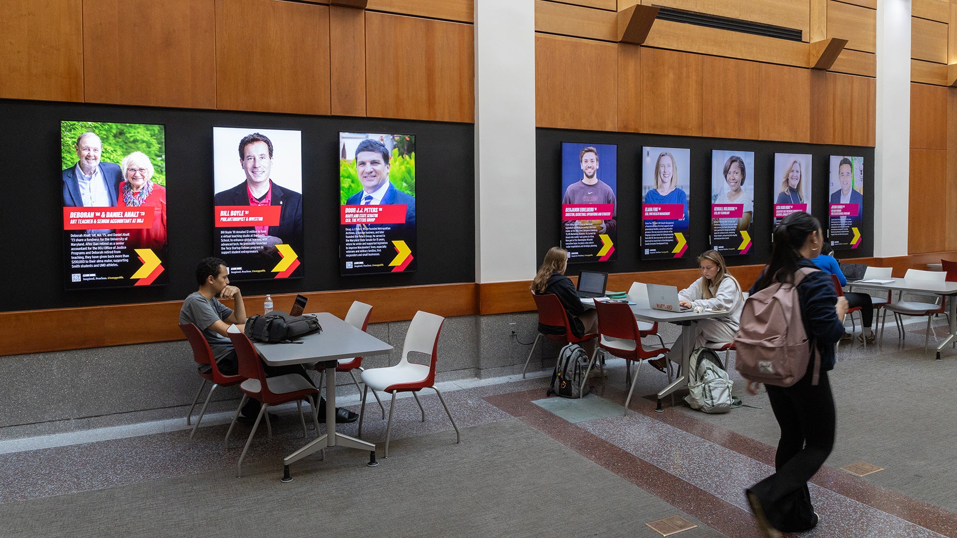 LED screens in Van Munching Hall display alumni profiles as students study and walk by.