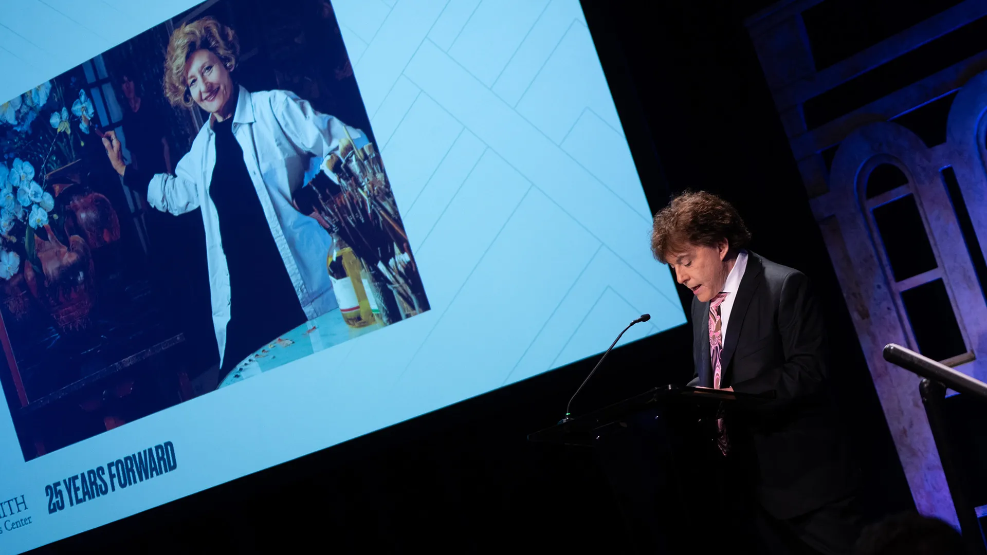 David Bruce Smith speaks at a podium during a University of Maryland event, with a large image of artist Clarice Smith displayed on a screen behind him.