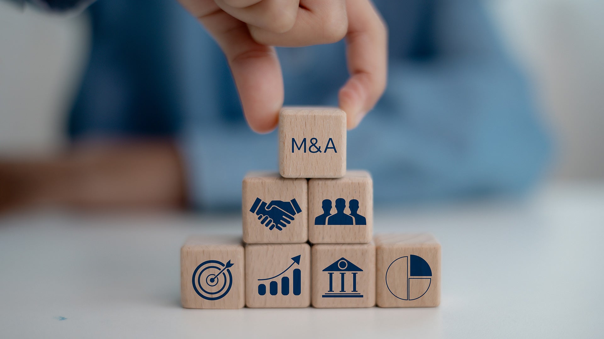 Hand placing a block labeled “M&A” atop stacked wooden blocks displaying business, finance and teamwork symbols.