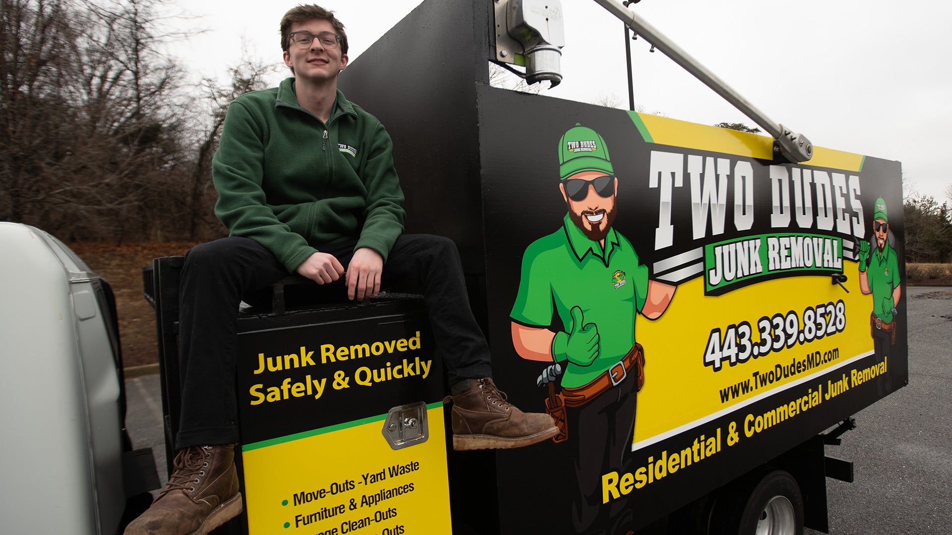 College student sits on the back of a branded “Two Dudes Junk Removal” truck, posing beside the company’s colorful logo and contact information.