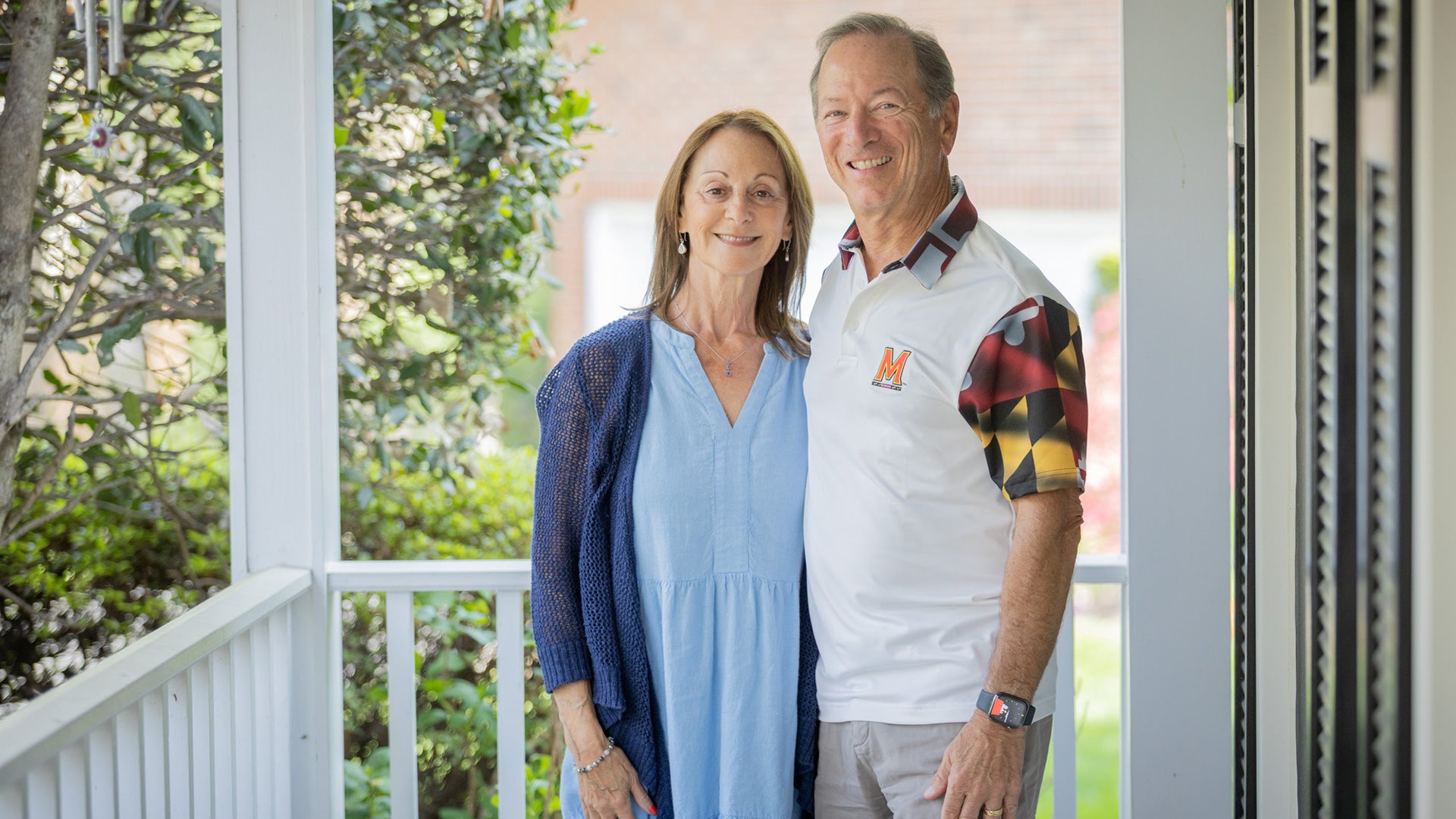 Craig and Lisa Adler stand together on a porch, smiling, with greenery in the background; he wears a University of Maryland shirt.