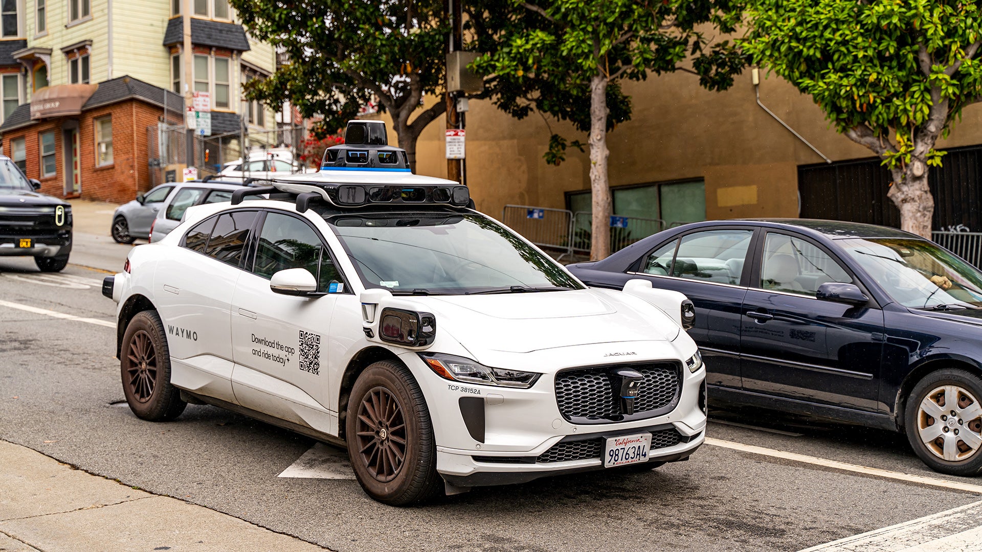 Waymo autonomous vehicle drives on a city street equipped with rooftop sensors and cameras.