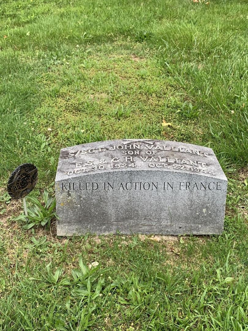 A gray granite military headstone set in grass, engraved with the name “S/Sgt. John Valliant, son of F.S. & C.H. Valliant, Oct. 30, 1921 – Oct. 12, 1944,” and the words “Killed in action in France.” A small bronze military medallion is placed in the ground beside the marker.