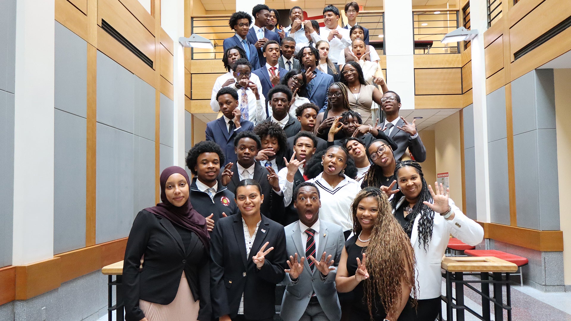A large group of young adults in business attire pose playfully together on an indoor staircase in a modern building.